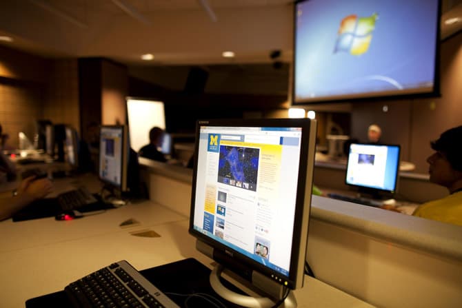 A computer screen in a darkened room full of computers, with a projector screen on in the background