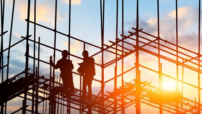 Two people in safety vests and hard hats stand on scaffolding in front of a sunset